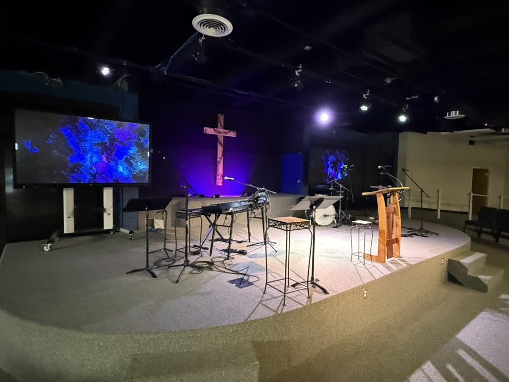 Curved stage from the side, with keyboard, drums, pulpit and lit cross